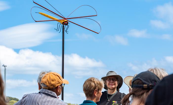 A picture of a tour guide leading a tour group next to a sculpture of a giant dragonfly.